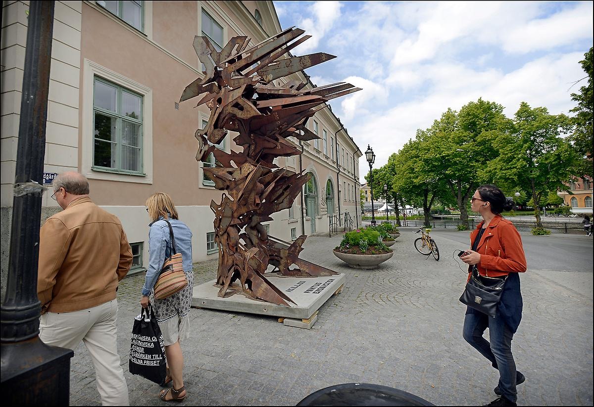 Utomhus på stan i Örebro går en kvinna intresserat förbi en skulptur som hon inte kan slita blicken ifrån. Skulpturen är rostfärgad och liknar en robot.
