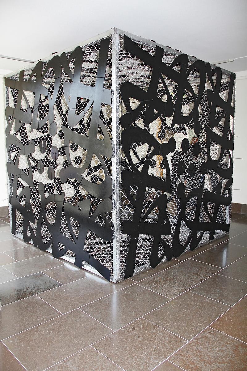 I ett rum står en bur i aluminium färgat stängsel. Buren pryds även av svarta former i olika storlekar, formerna liknar nästan bokstäver. 
