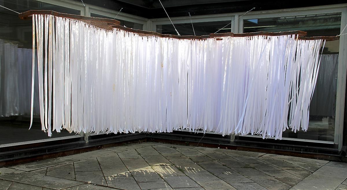 I luften hänger en träram i en avlång böljande form och ner från den hänger långa vita pappersremsor. 