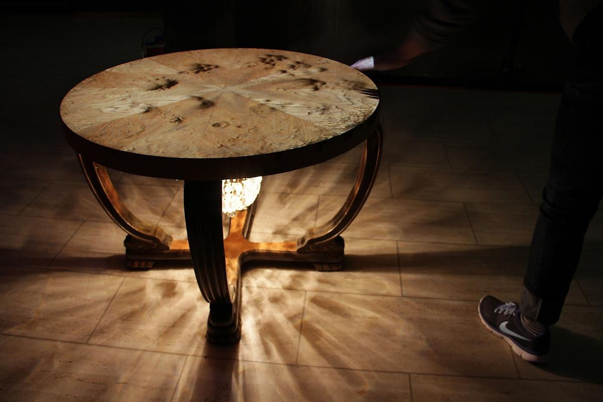 Inomhus på ett golv står ett runt träbord. Under bordet sitter en lampa som skapar mönster på golvet runt bordet. 