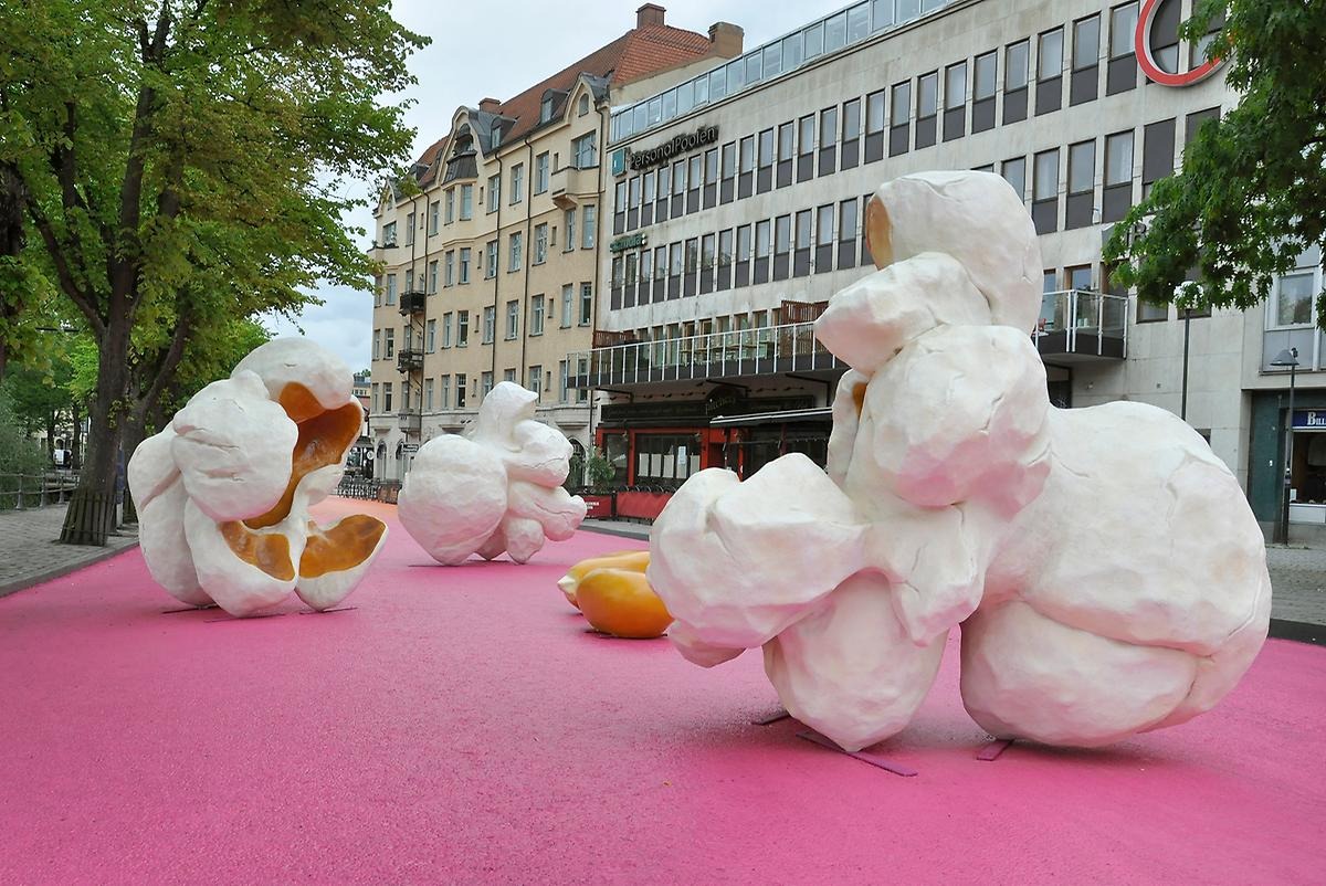 På en rosamålad gata står tre överdimensionerade popcorn i verklighetstrogna färger och former. Popcornen är formade så det går att sitta i dem och på den rosa gatan ligger även två överdimensionerade majskorn.