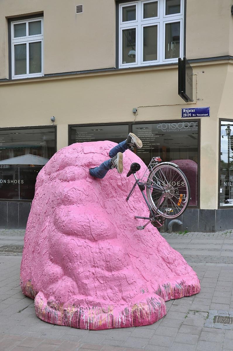 På marken står en stor rosa skulptur formad som en snödriva. Ur skulpturen sticker en halv cykel ut och två ben som har jeans och gympaskor på sig. 