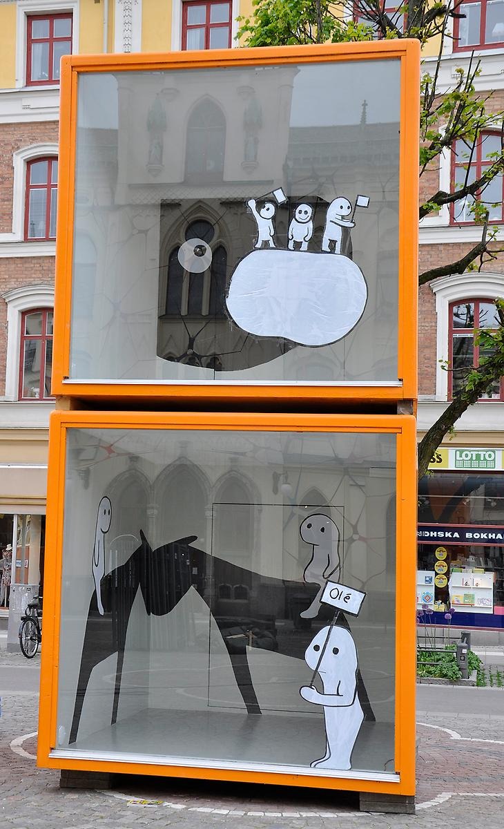 Utomhus på Stortorget i Örebro står två orangea glasmontrar. I dem beskådas en kort ritad serie då montrarna innehåller olika små gubbar i utskurna bläckteckningar.