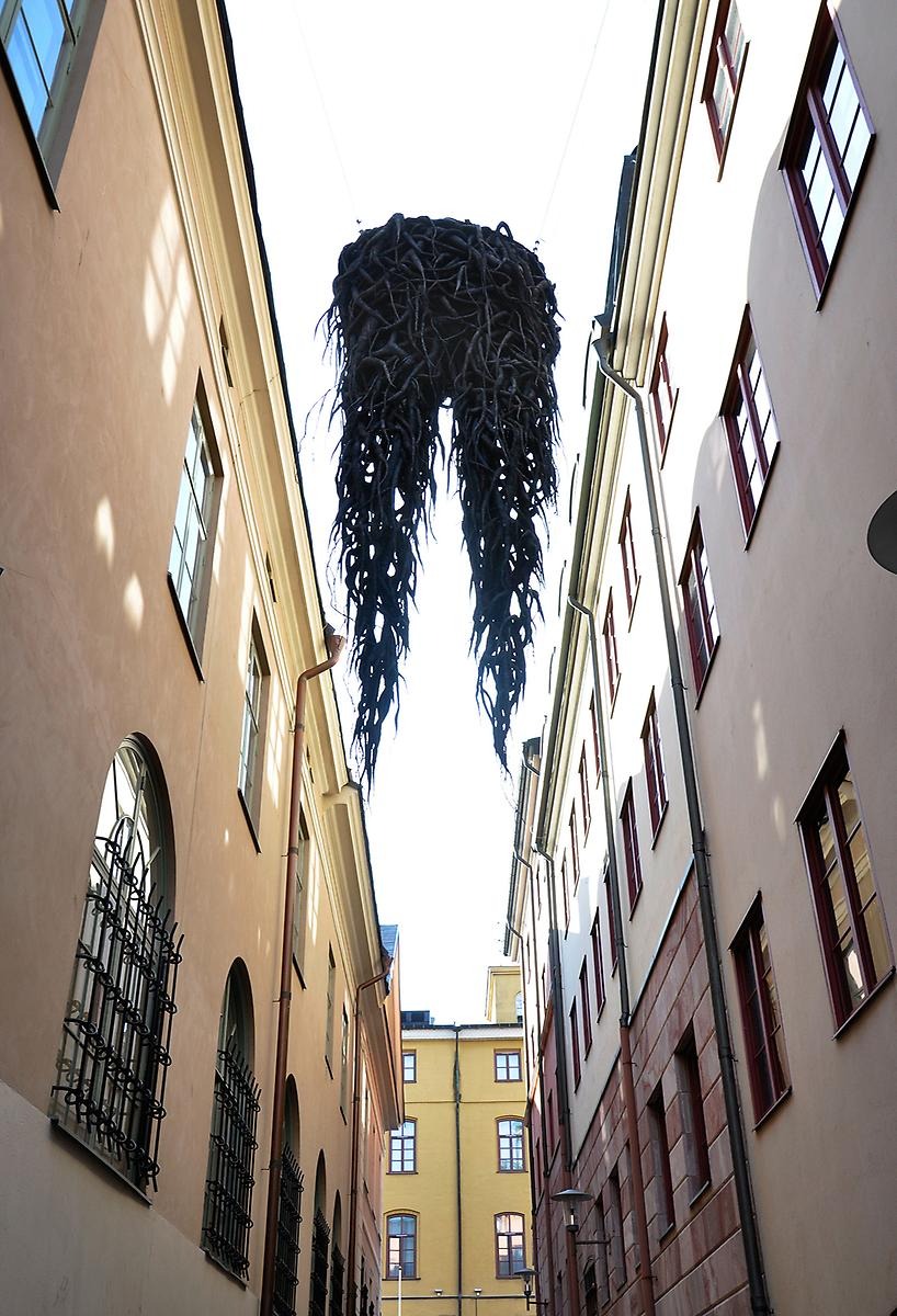 På en smal gata mellan två höga husbyggnader hänger en svart skulptur i vaxad textil.