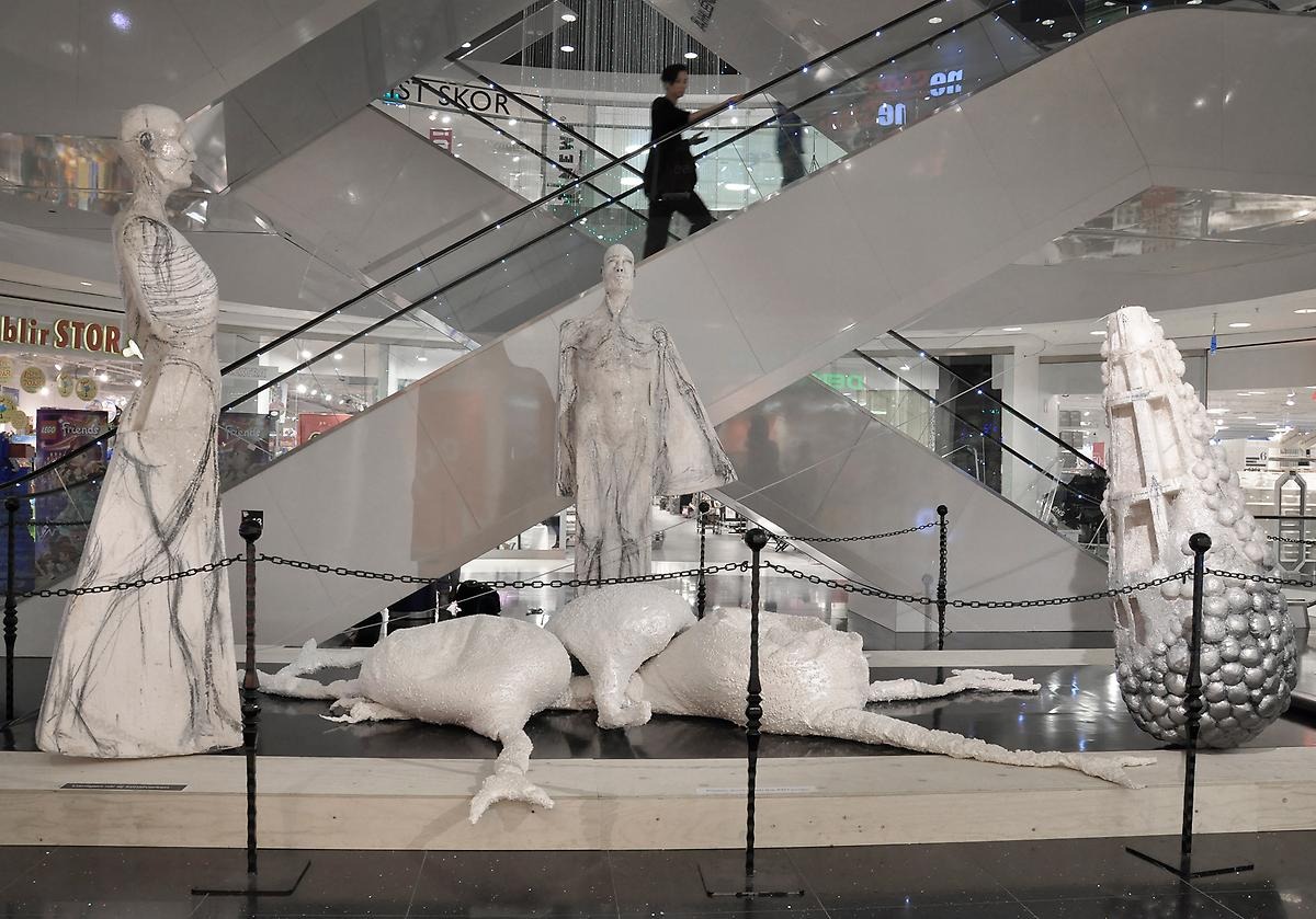 På bottenvåningen av gallerian Vågen står en installation av fyra vita, stora skulpturer och i bakgrunden åker en kvinna ner för gallerians rulltrappa. Installationen består av två långa och mäktiga människoskulpturer.
