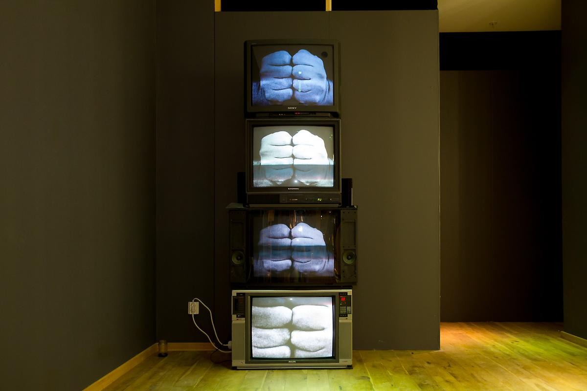 I ett mörkt rum med gråväggar står fyra stycken analoga TV-apparater staplade på varandra. TV-apparater visar närbilder på två knutna nävar mot varandra.