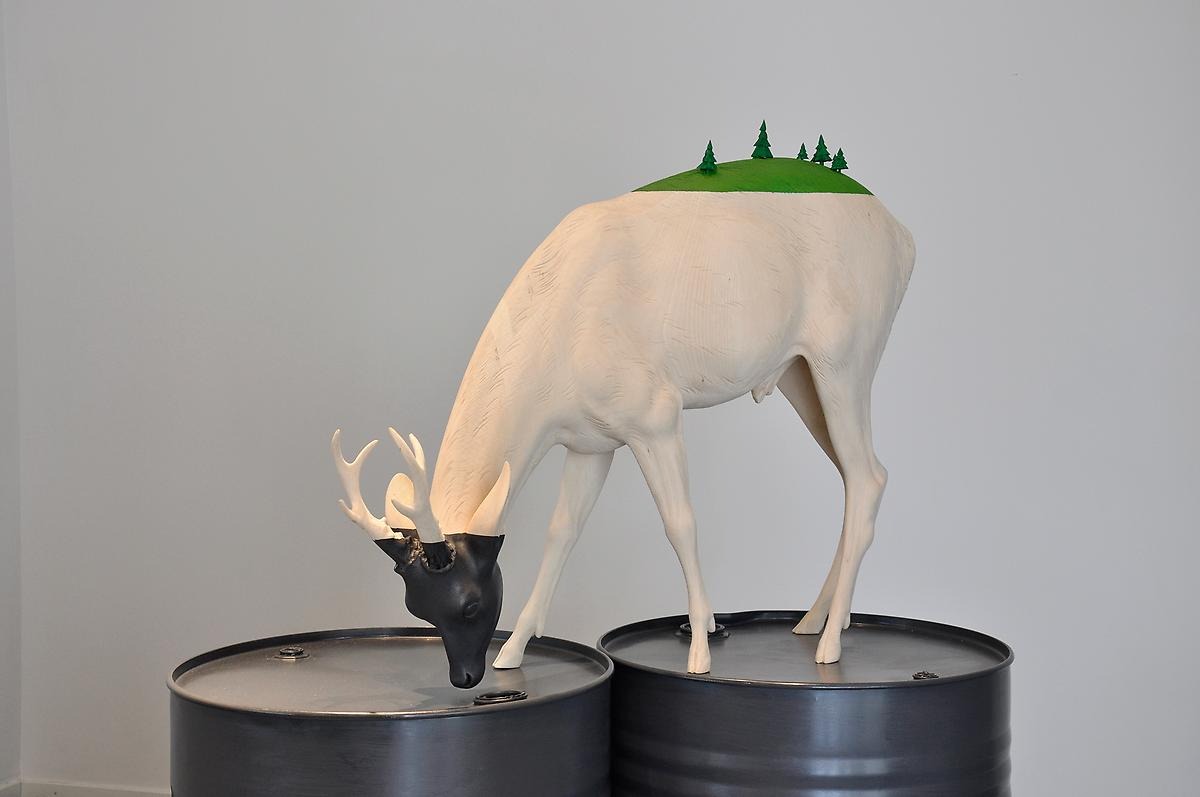 En skulptur av ett vit rådjur står på två oljefat. Rådjurets huvud är svart målat och lutat neråt mot oljefatet samt rådjurets rygg är grönmålad och har små trä skulpturer ståendes på sig.