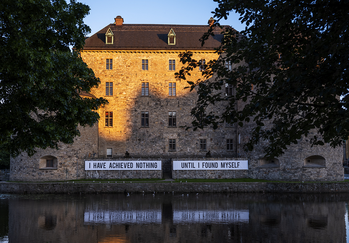 På Örebro slott sitter två stora svartvita banderoller Med texten "I HAVE ACHIEVED NOTHING UNTIL I FOUND MYSELF" uppsatta på nedre delen av fasaden. I det mörka vattnet nedanför syns banderollernas reflektioner. Ovanför syns lövverken från två träd.