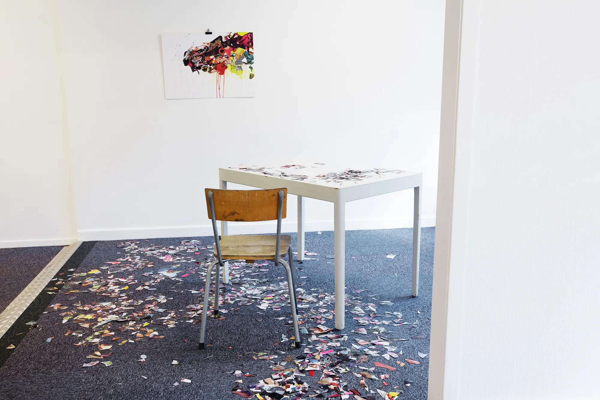 I ett rum syns en abstrakt färgglad tavla upphängd på en vit vägg. Snett framför tavlan står ett vitt bord och en stol. Utspritt på bordet och golvet ligger en stor mängd små pappersbitar som har samma färger som tavlan.