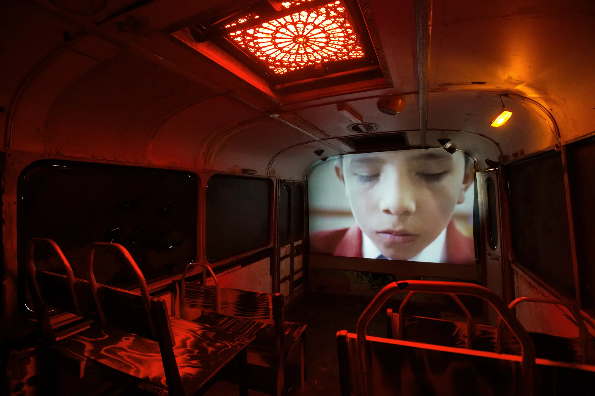 Bilden är tagen inuti bussen där belysningen är röd. Längst fram är en projektorskärm som visar upp en när bild på en pojke som blundar.