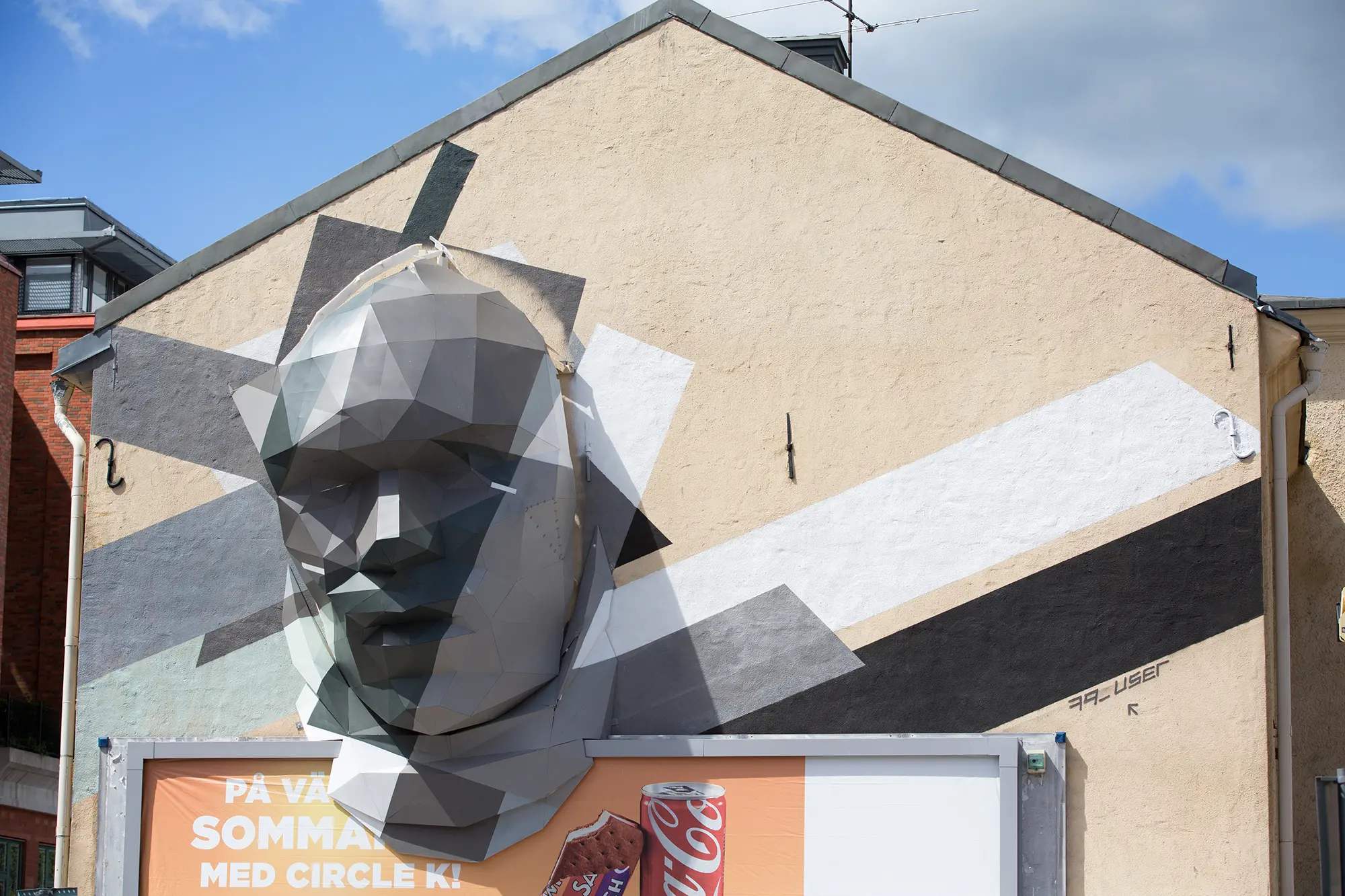 På en en husfasad ovanför en reklamplansch sitter en stor grå skulptur föreställande ett ansikte uppsatt. Ansiktet är uppbyggt av många plana ytor som sitter ihop på ett kantigt sätt. I bakgrunden syns olika målade färgfält svart, vitt och grått.