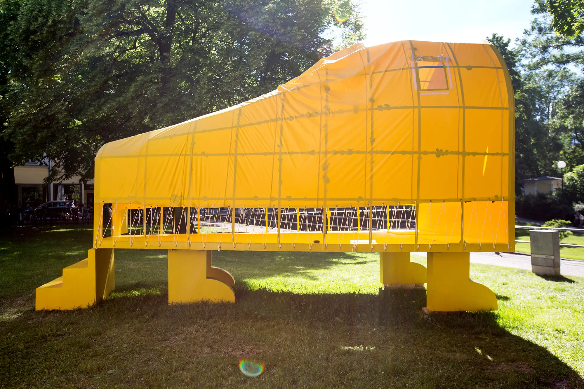 På gräsmattan i en park står en stor gul skulptur i formen av ett avlångt tält. Skulpturen är upphöjd på fyra gula ben.  Taket är högre på ena kortsidan och lutar ned mot en ingång på den andra sidan. Framför ingången står en gul trappa.