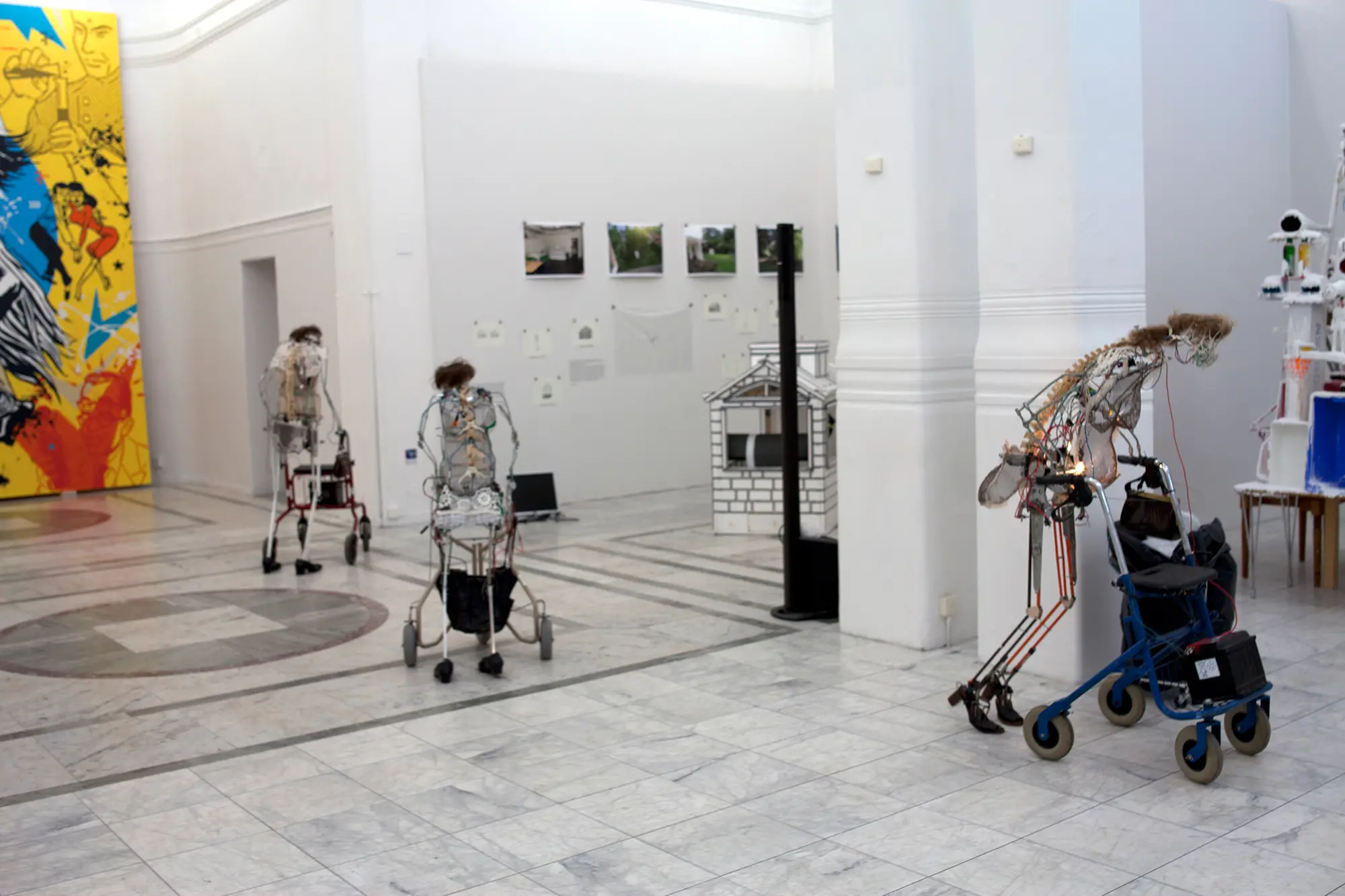 I ett stort ljust rum syns en installation bestående av tre människolika robotar som står framåtlutade ovanför varsin rollator med batterier på. De är uppbyggda av ett metallskelett  och sladdar.