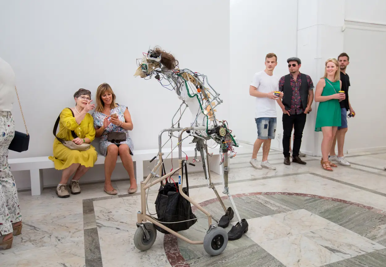 I en utställningssal med syns en installation bestående av en människolik robotskulptur framåtlutad över en rollator. Skulpturen är uppbyggd av tunna metallrör, sladdar, glasögon och hår. I bakgrunden syns flera människor som betraktar skulpturen.