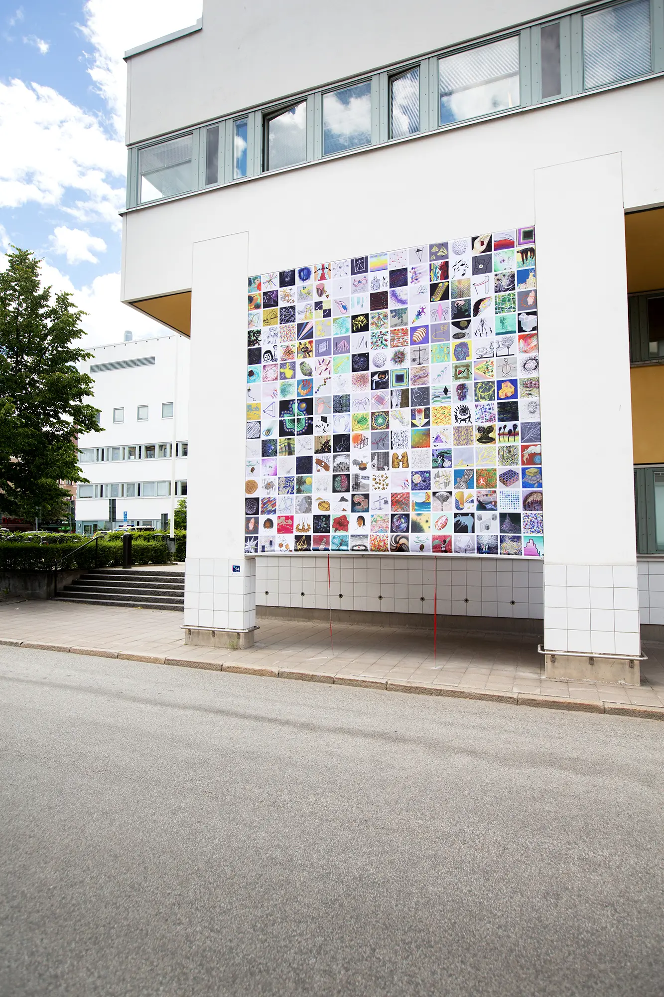 På en vit byggnad sitter en väggmålning upphängd mellan två pelare. Målningen består av en stor mängd små kvadratiska bilder i rader. De små bilderna är både föreställande och abstrakta i olika stilar och färger.