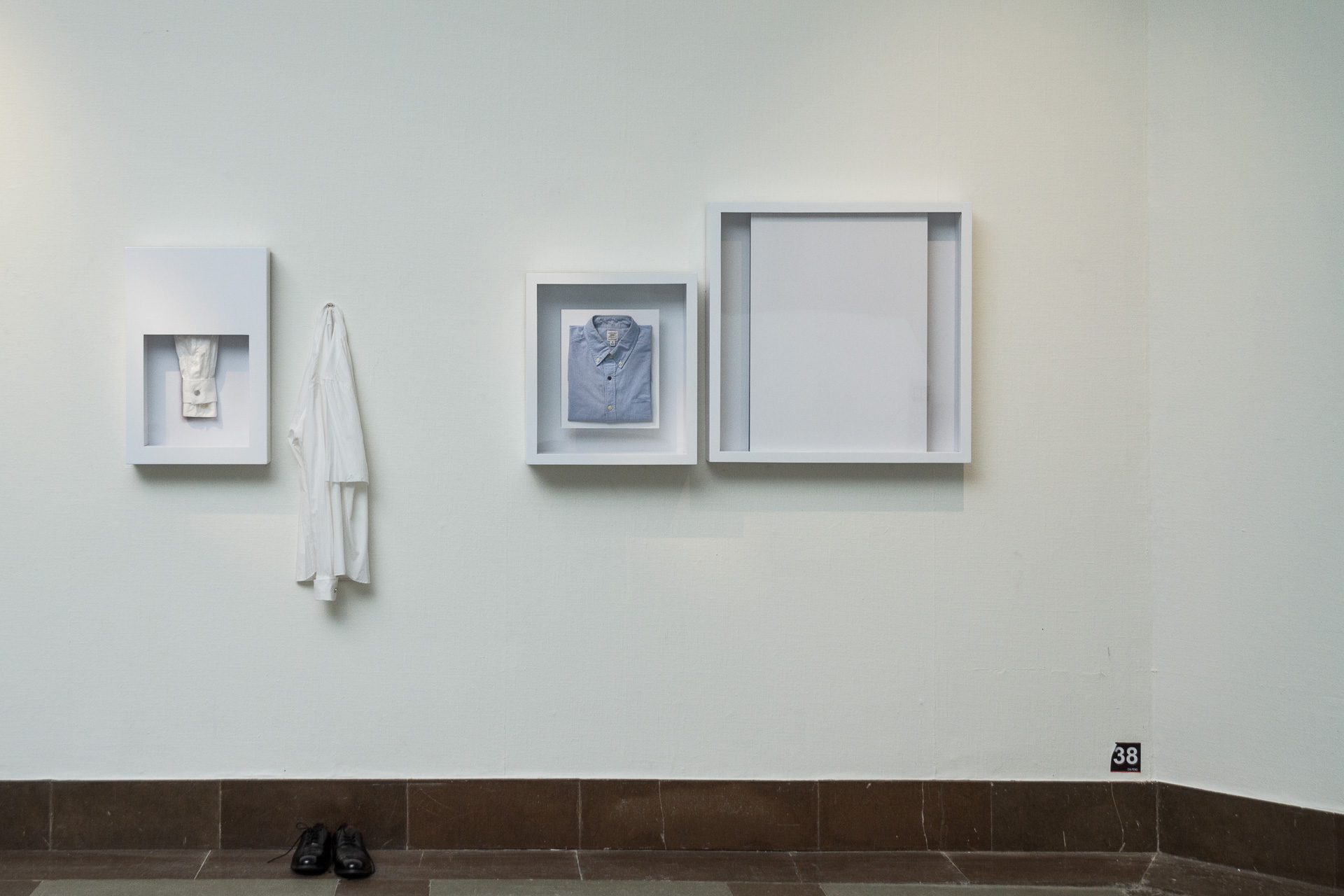 På en vit väg hänger en installation som innehåller tre olika ramar och en upphängd T-shirt. Ur första ramen kikar en skjortärm ut, i den andra ramen ligger en bild på en vikt ljusblå skjorta och sista ramen innehåller en vit bild.