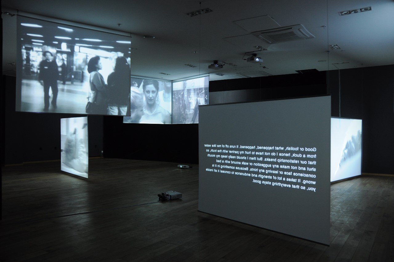 I ett mörkt rum står sex olika projektordukar som spelar upp olika svart vita bilder på människor. Tre av projektdukarna hänger i taket och tre står på golvet. 