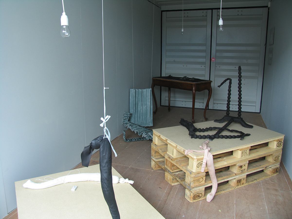 I en container står tre EU-pallar radade på varandra även ett träbord står på golvet och i taket hänger lampsladdar. 