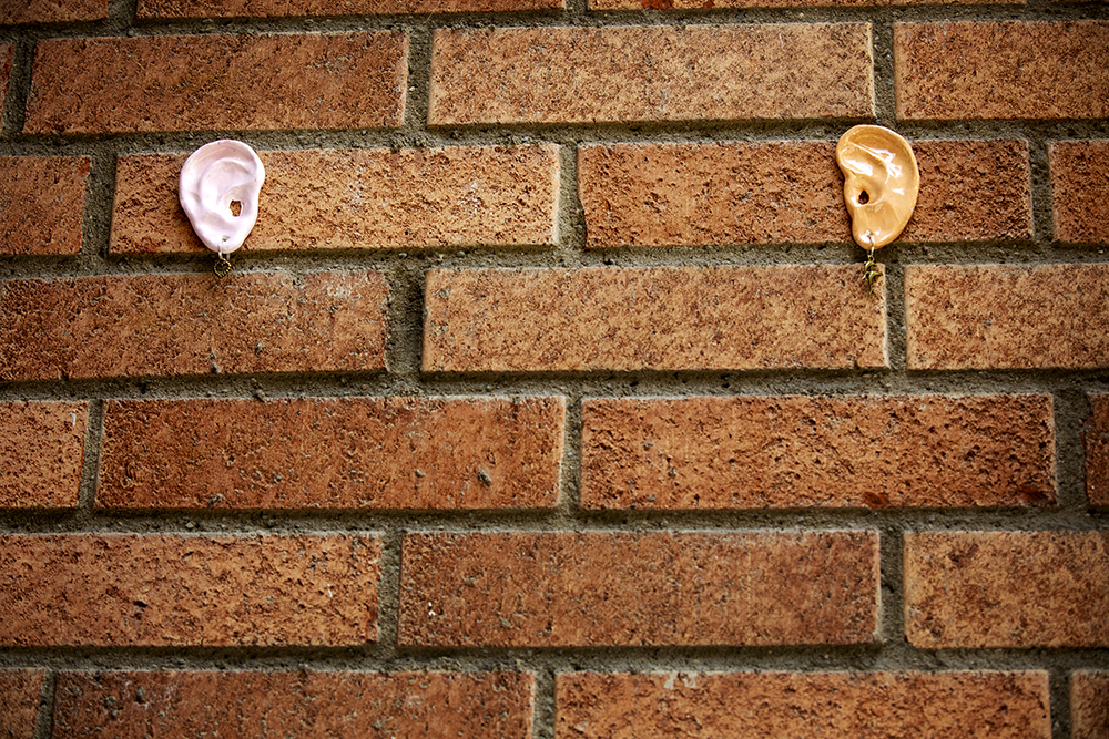 Two ceramic ears with earrings are mounted on an orange brick wall.