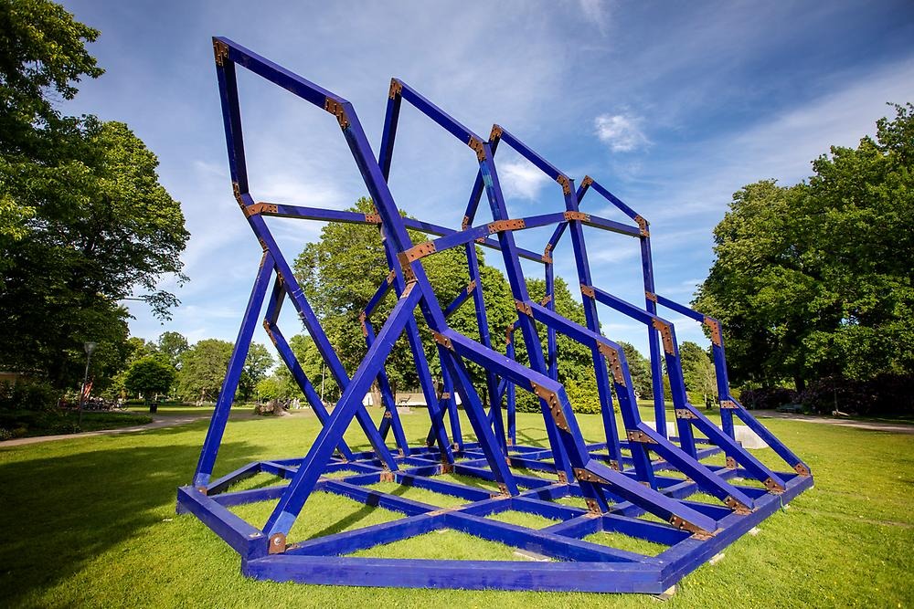 A construction made of blue painted wood, it resembles a church in construction, but there are no walls. It is located in a park environment.