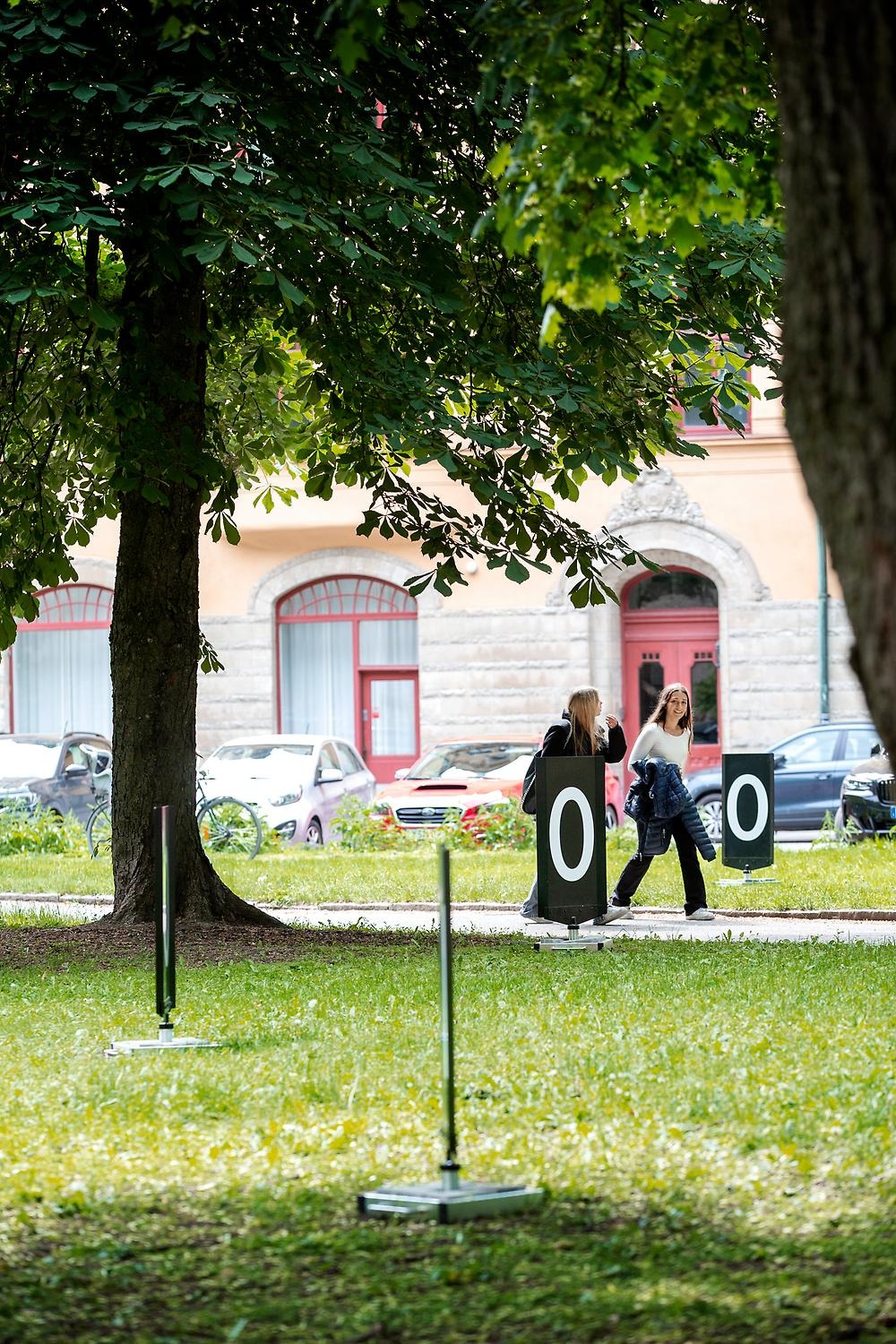 In a park, black pavement signs with the numbers 1 and 0 are placed. The picture shows four signs and two people walking by in the background.