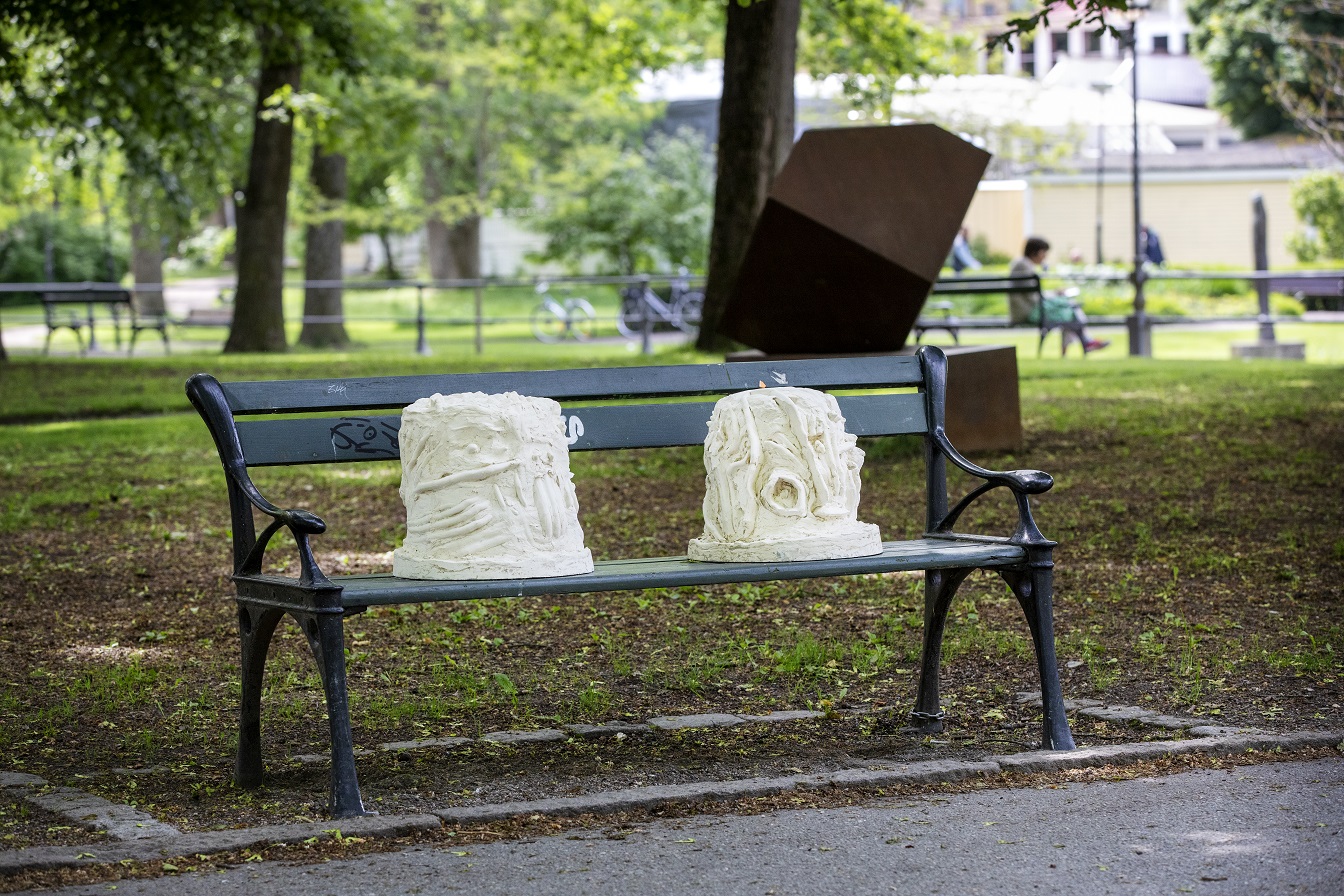 Outside on a park bench with a park environment behind it sit two white and yellow sculptures. In the sculptures, you can make out hands that converse in sign language with each other.