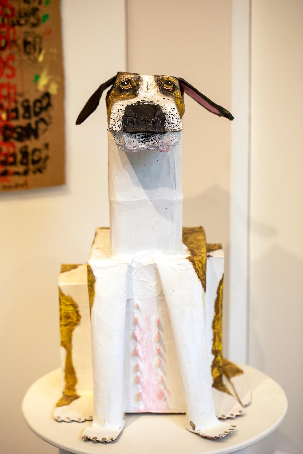 En skulptur. Skulpturen föreställer en hund. Den är gjord av toarullar, kartong och är målad vit och brun. 