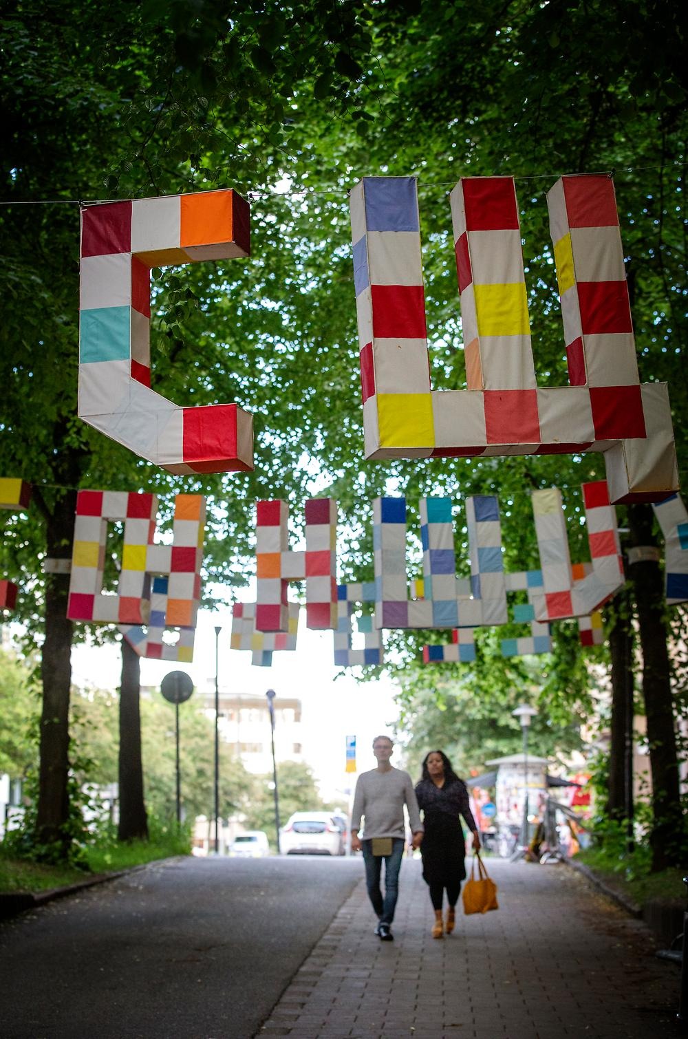 Above a bicycle lane in the trees hang large letters from the Latin, Hebrew and Cyrillic alphabets. The letters are three-dimensional, white and with sewn-on rectangular textile pieces in blue, red and orange tones. Underneath the letters two people can be seen walking and holding hands.