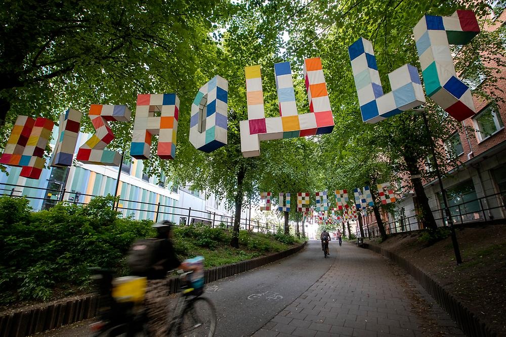 Above a bicycle lane in the trees hang large letters from the Latin, Hebrew and Cyrillic alphabets. The letters are three-dimensional, white and with sewn-on rectangular textile pieces in blue, red and orange tones.