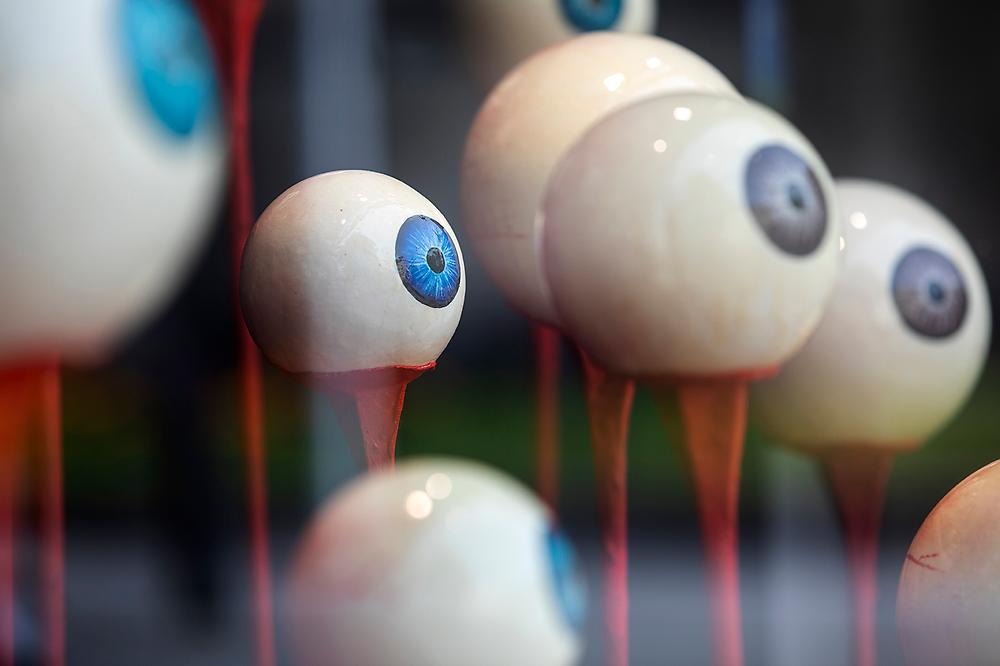 Close-up of large staring eyes without eyelids in different sizes sitting on each red stick, the iris colors of the eyes are blue and brown. The artwork is placed in a shop window, which means that it is reflected in the glass.