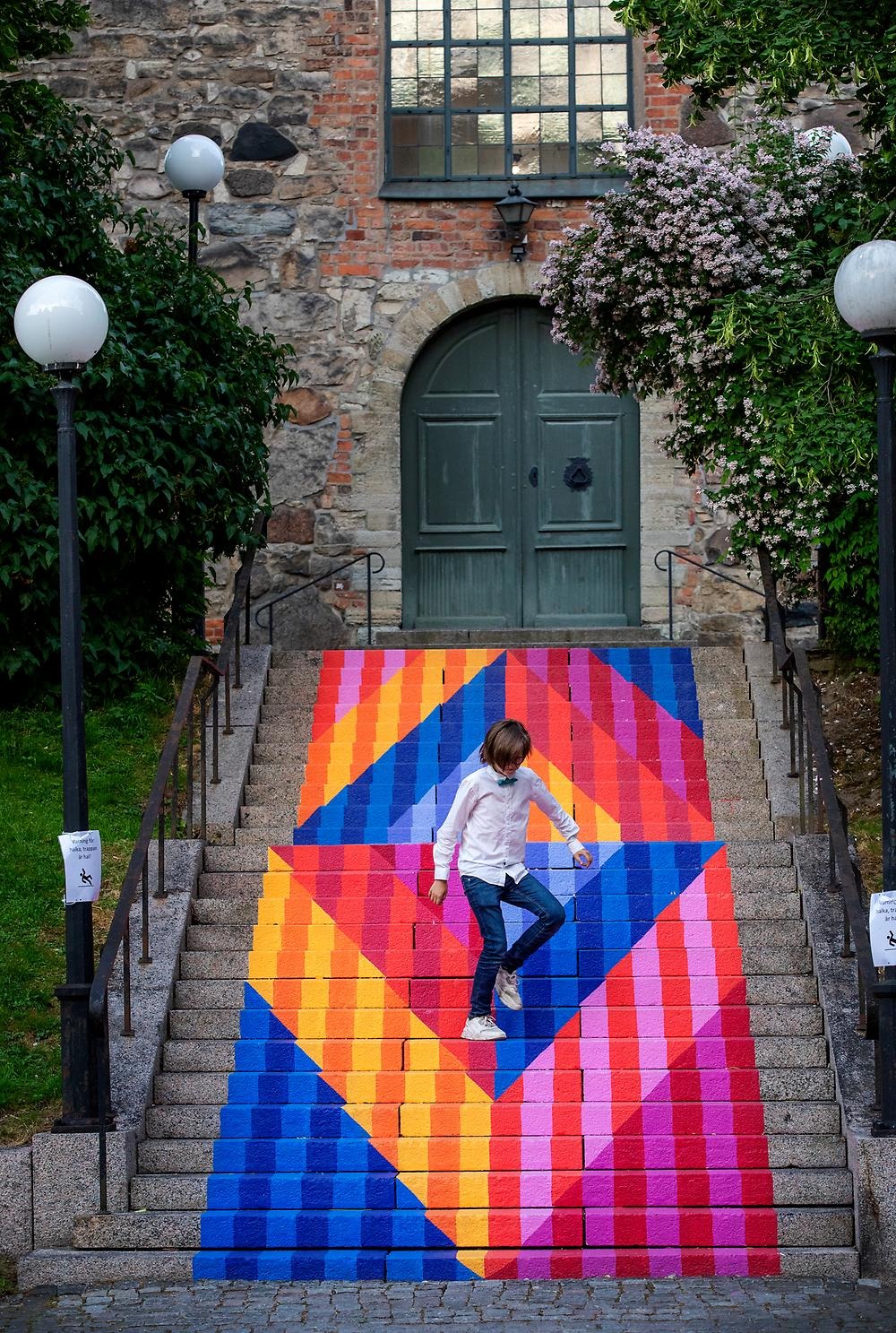 The stairs up to St. Nicholas Church are illuminated by a colorful work of art in different geometric shapes that carry the steps between different ages. You can't help but be happy when you see this fantastic staircase! In the background you can see St. Nicholas Church, green trees and a boy with a shirt and bow tie who is jumping around the stairs.