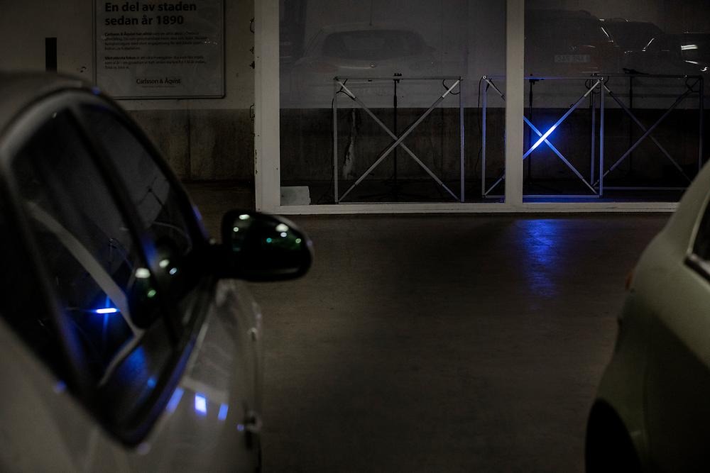 In the foreground are the sides of two parked cars. Behind the cars you can see the room where the installation is. The square modules with crosses in them are currently off, but you can sense a blue glow. Otherwise, the light in the image is dark.