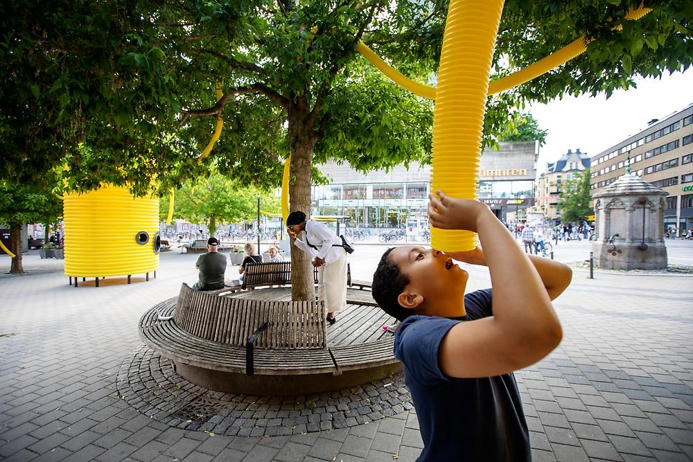Two people are engaged in talking to each other through yellow drainage pipes. They are moving in a square environment and one of them has stepped onto a bench.