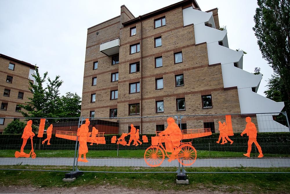 A high fence follows the promenade by the water towards the city park. The fence consists of orange embroidery depicting people in motion. The people cycle, walk and ride electric scooters.