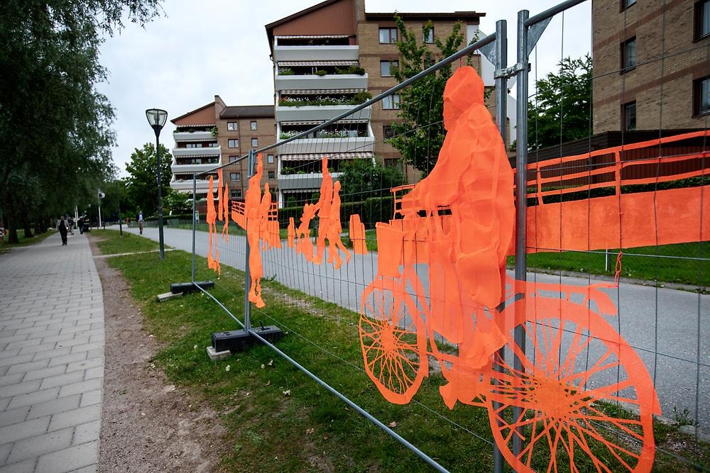 A high fence follows the promenade by the water towards the city park. The fence consists of orange embroidery depicting people in motion. The people cycle, walk and ride electric scooters.