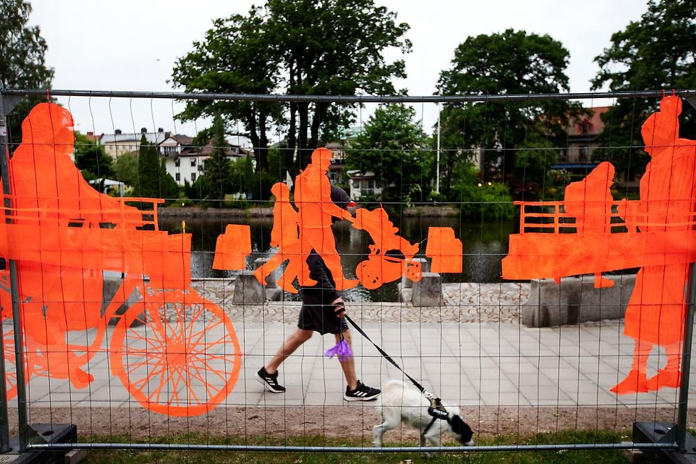 A close-up of the high fence that follows the promenade by the water towards the city park. The fence consists of orange embroidery depicting people in motion. The people cycle, walk and ride electric scooters. In the background, a person walks his dog.