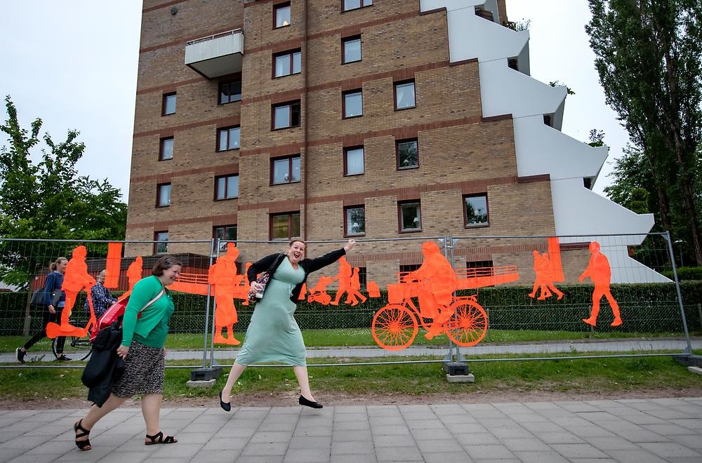 Two women laugh and one jumps in front of the large fence that follows the promenade by the water towards the city park. The fence consists of orange embroidery depicting people in motion. The people cycle, walk and ride electric scooters.