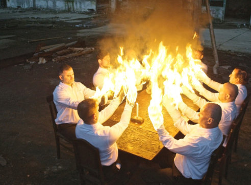 The image is taken from a video artwork where eight men dressed in white sit around a table. They stretch out their arms that burn halfway down to the elbows.