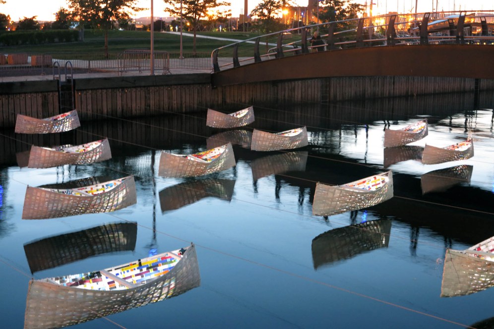 It is evening and eleven canoe-like shapes can be seen hovers above the water's surface. The inside of the canoes is decorated with a pattern of colorful squares and the inside is illuminated in the dark.