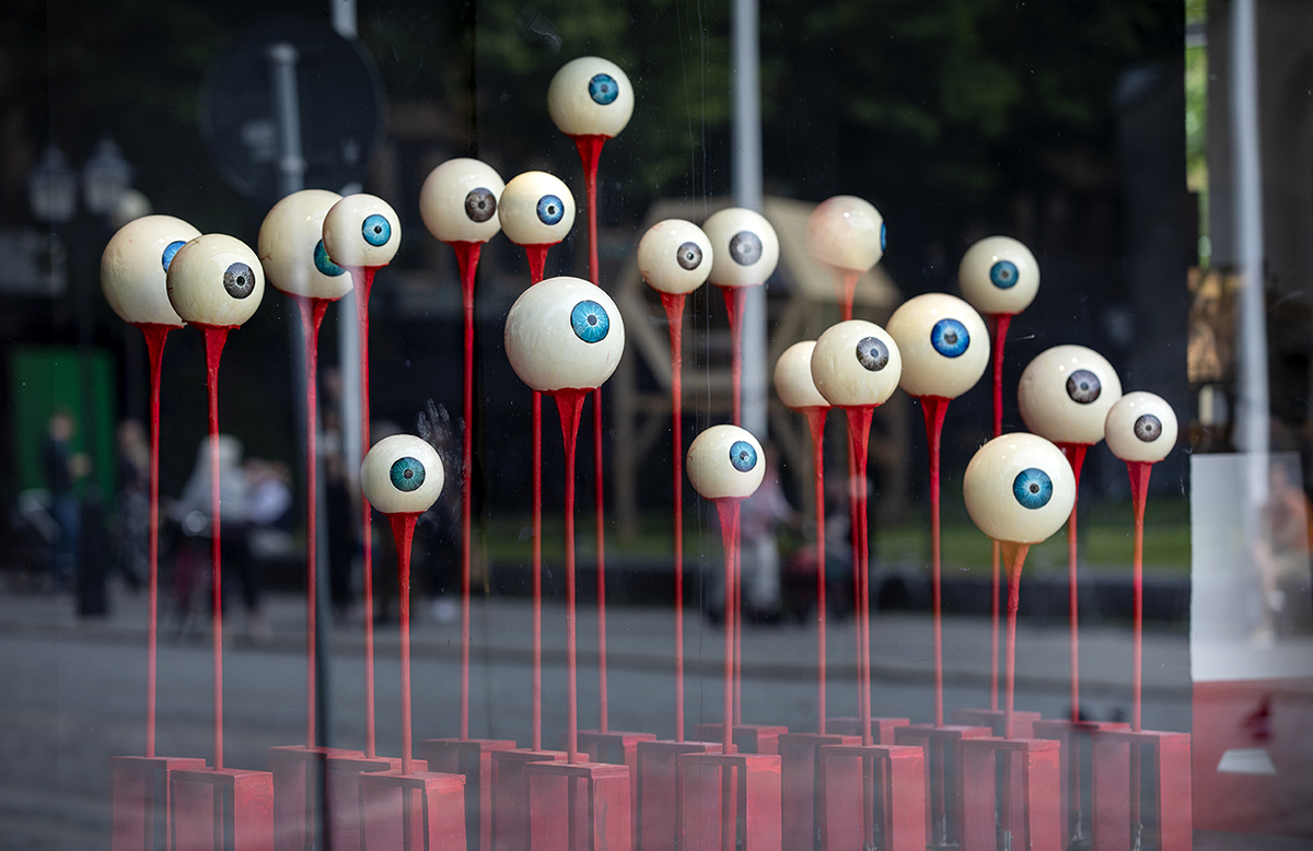 20 large staring eyes without eyelids in different sizes sit on each red stick, the iris colors of the eyes are blue and brown. The artwork is placed in a shop window, which means that it is reflected in the glass.